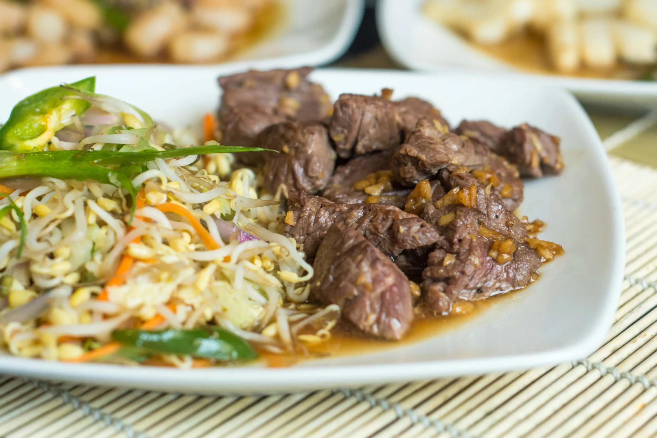 A plate of stir-fried beef and bean sprouts with a light brown sauce. The beef is cut into small pieces and is in the foreground of the image, while the bean sprouts are in the background. The plate is on a bamboo placemat.