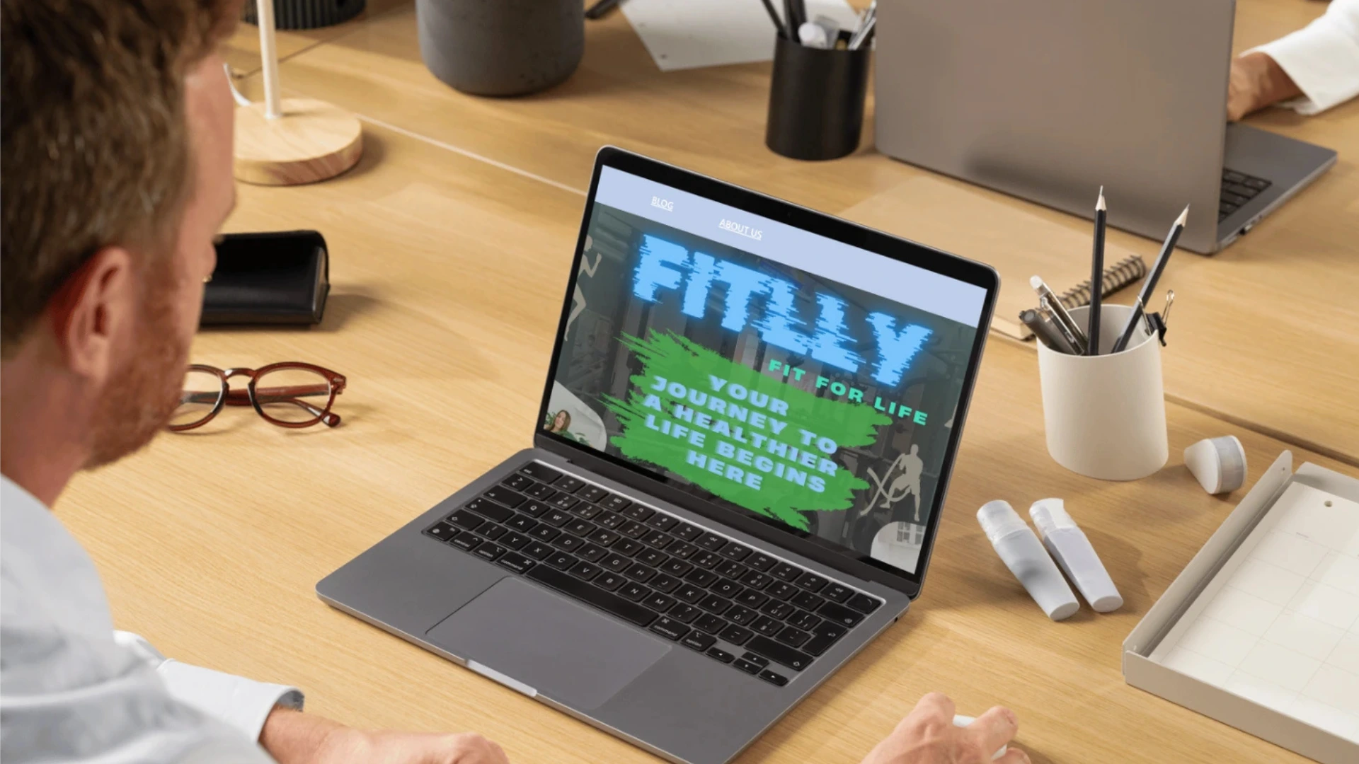 A person interacting with a laptop displaying a fitness-related website titled "FITLLY: Fit for Life" with slogans promoting a healthier lifestyle.
